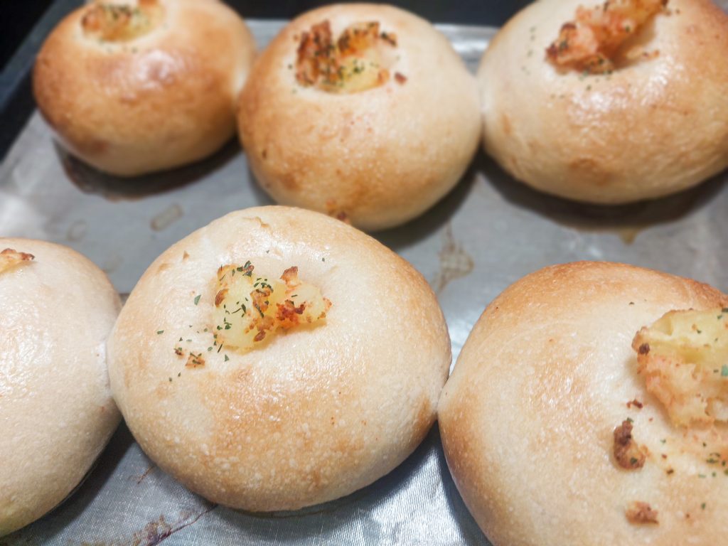 こまいぬベーグル堂のまんまる惣菜パン。ふっくら丸い形にチーズとハーブがトッピングされた焼き立てパンがトレーに並ぶ