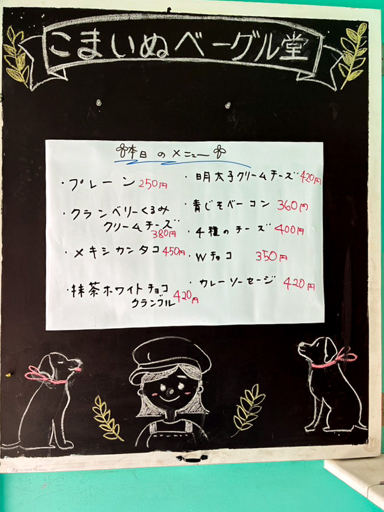 こまいぬベーグル堂の手書き黒板メニュー。プレーン250円・明太子クリームチーズ420円・クランベリーくるみクリームチーズ380円・青じそベーコン360円・4種のチーズ400円・メキシカンタコ450円・Wチョコ350円・抹茶ホワイトチョコクランブル420円・ウレーソーセージ420円。犬のかわいいイラスト入り