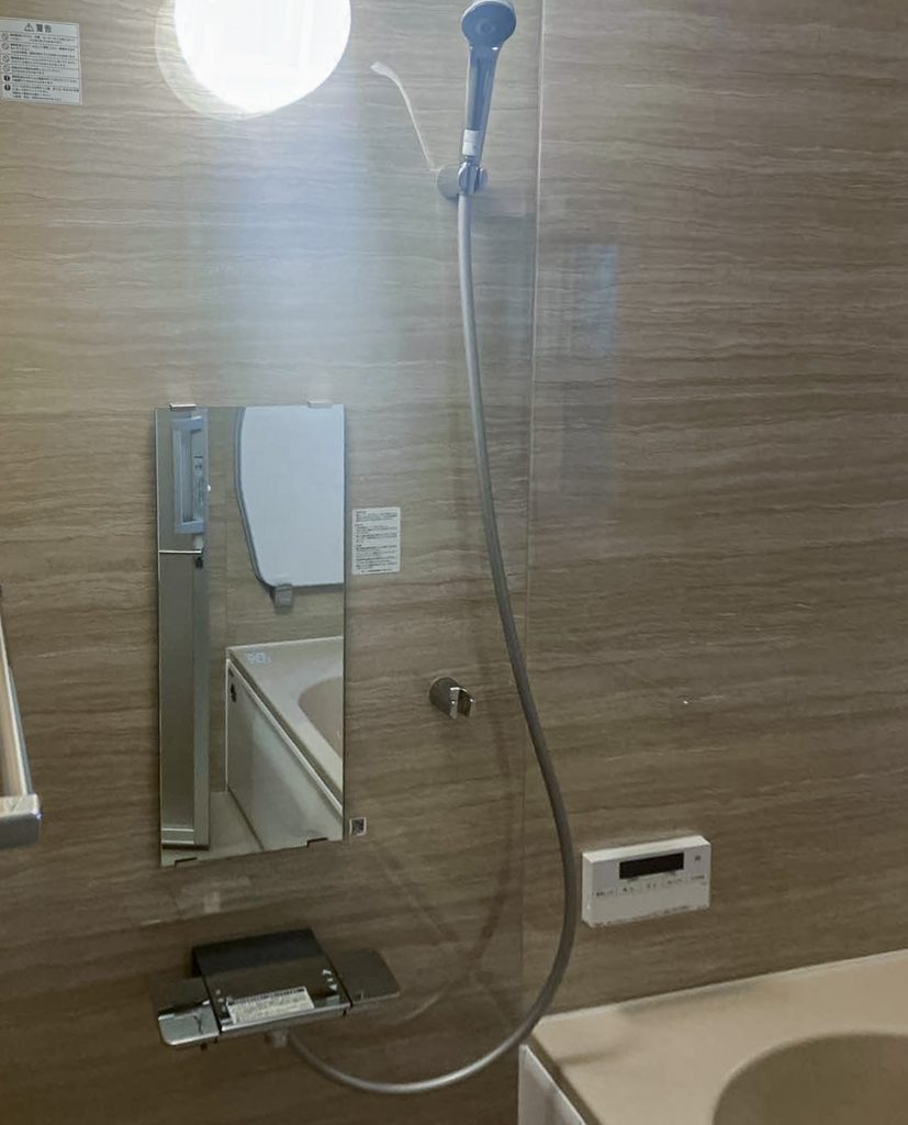 柏市M様邸の浴室リフォーム後のシャワーエリア。木目調の壁パネルにシャワー水栓・鏡・浴室暖房乾燥機リモコンが配置された清潔感のある空間